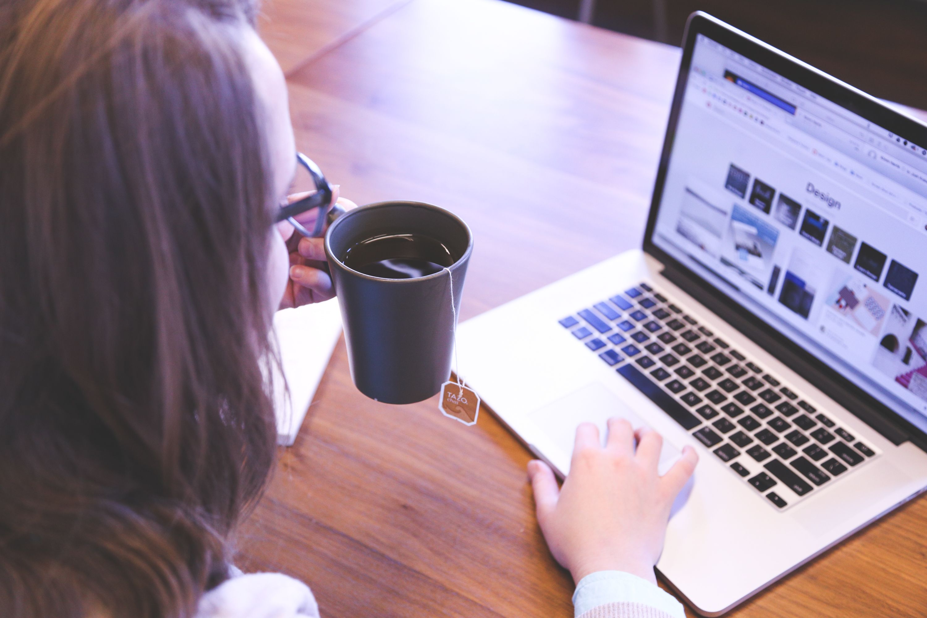 Woman with glasses drinking tea while browsing a design website on a laptop at a wooden table.