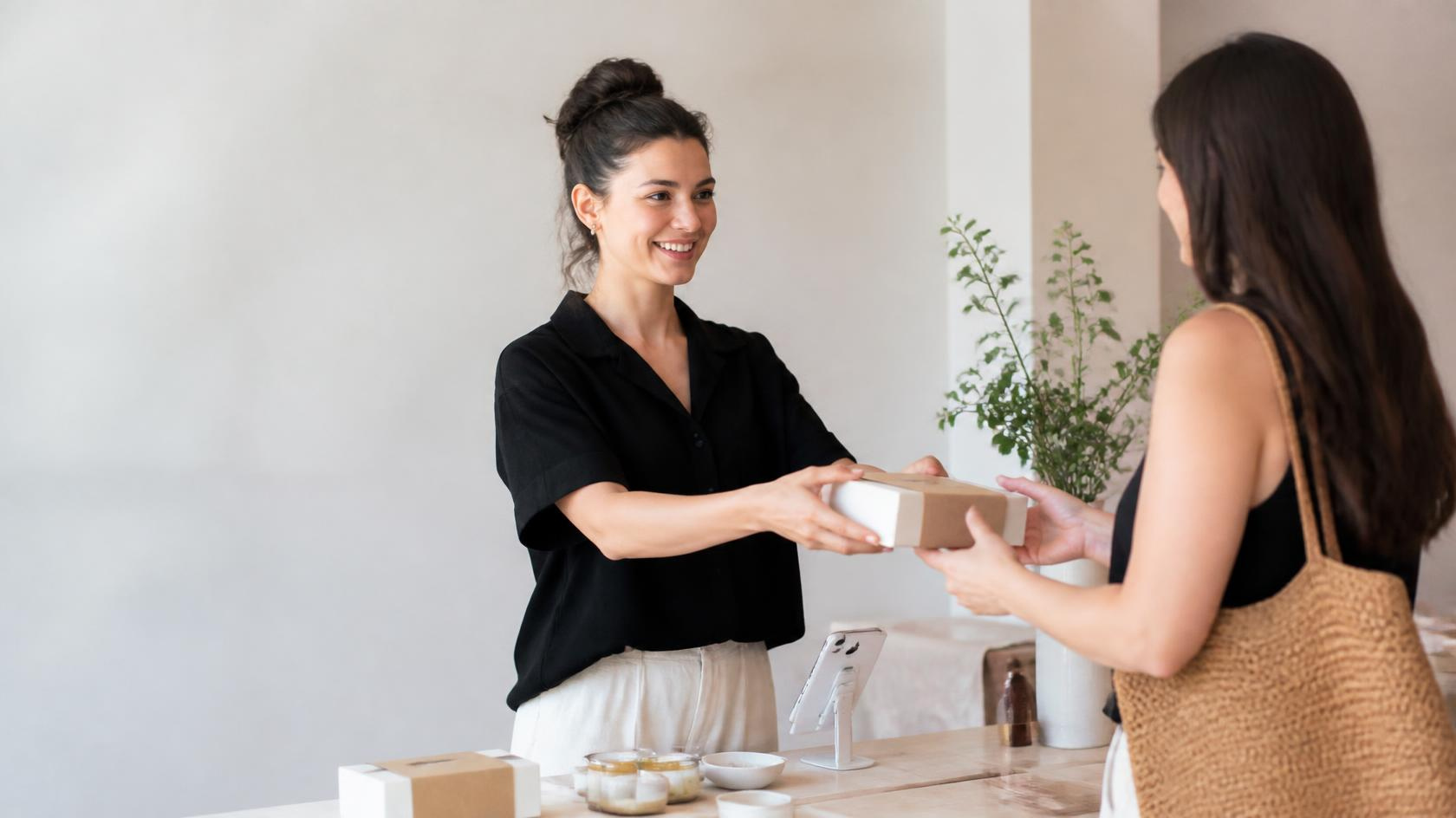 Founder handing a packaged product in a minimalist box to a smiling customer across a wooden table at a small pop-up stand.