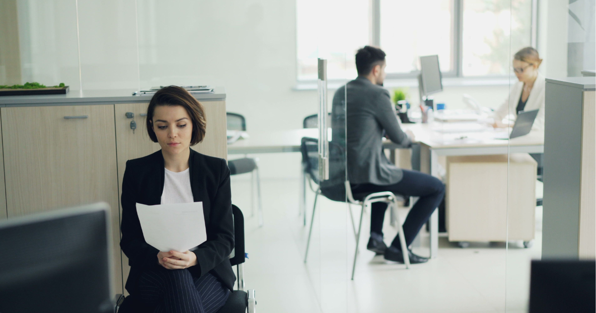 Young woman in a black blazer sits outside a glass-walled office, reading papers while two people work at desks inside.